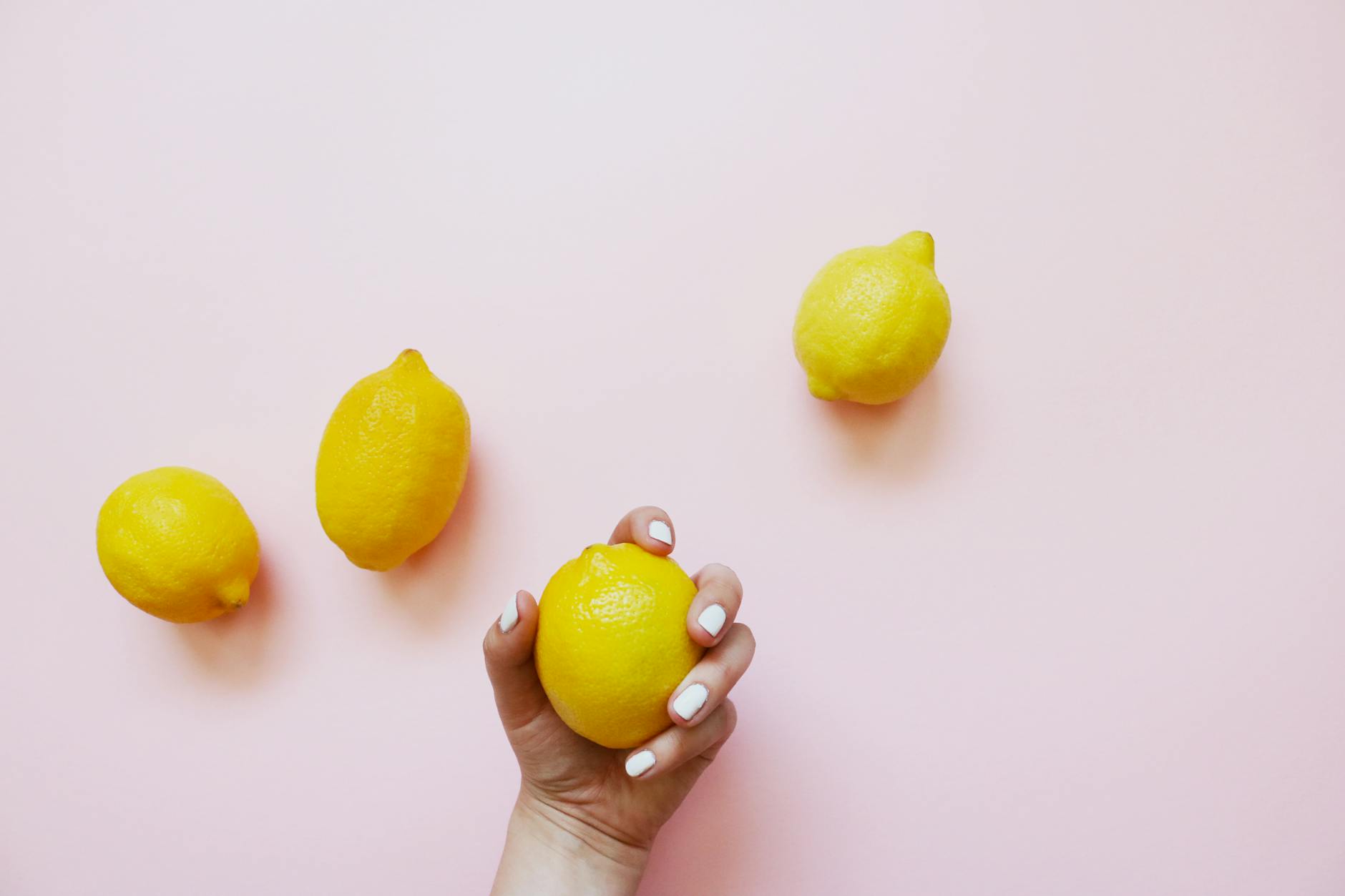 Una mano sosteniendo un limón fresco sobre un fondo rosa suave, rodeado de tres limones adicionales